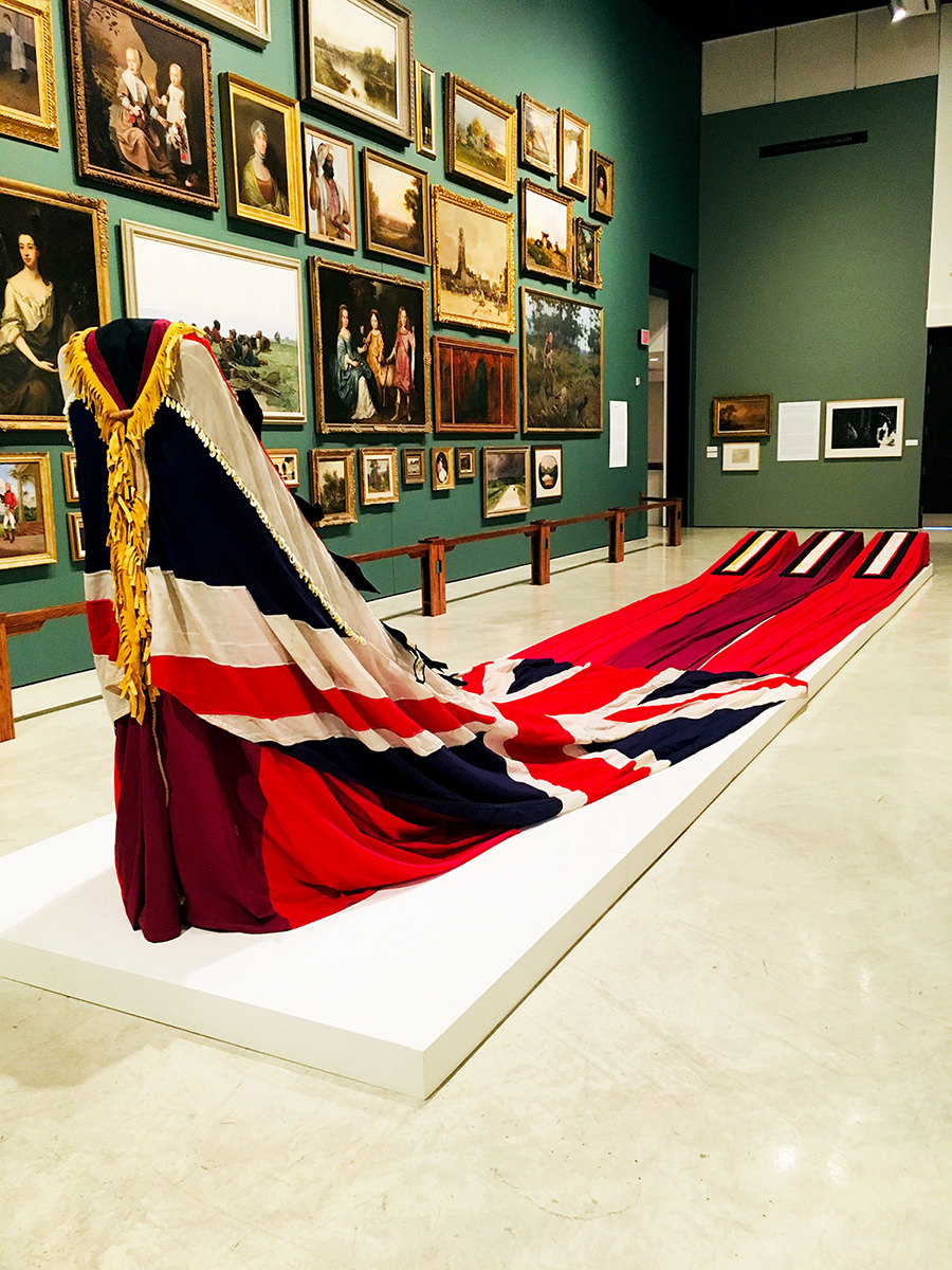 A red, white and black robe with a long train is displayed in front of a wall of paintings. 