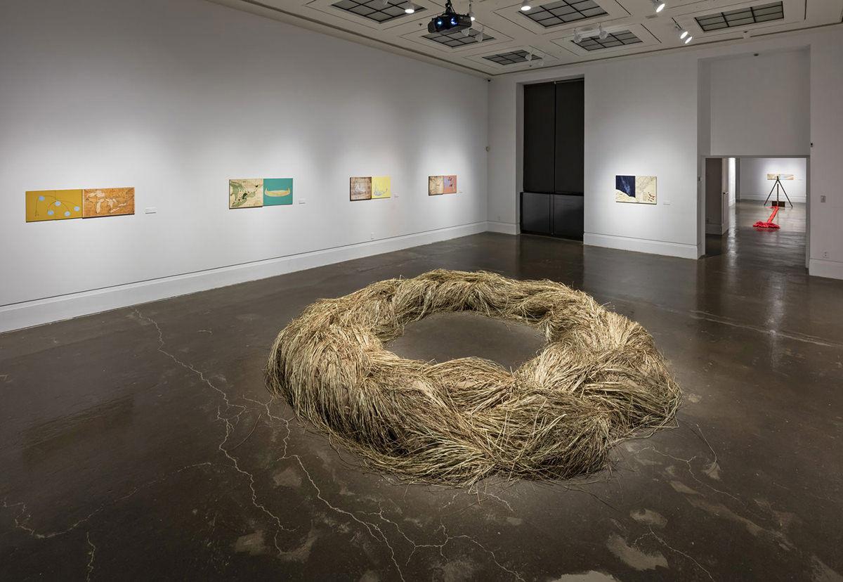 Various works of art are displayed on walls around a circle of reeds on the floor of an exhibition room. 