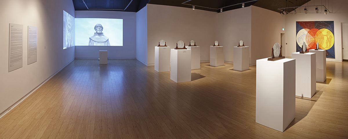 Glass discs in wooden holders sit on white stands next to a video projected on an exhibition room wall. 
