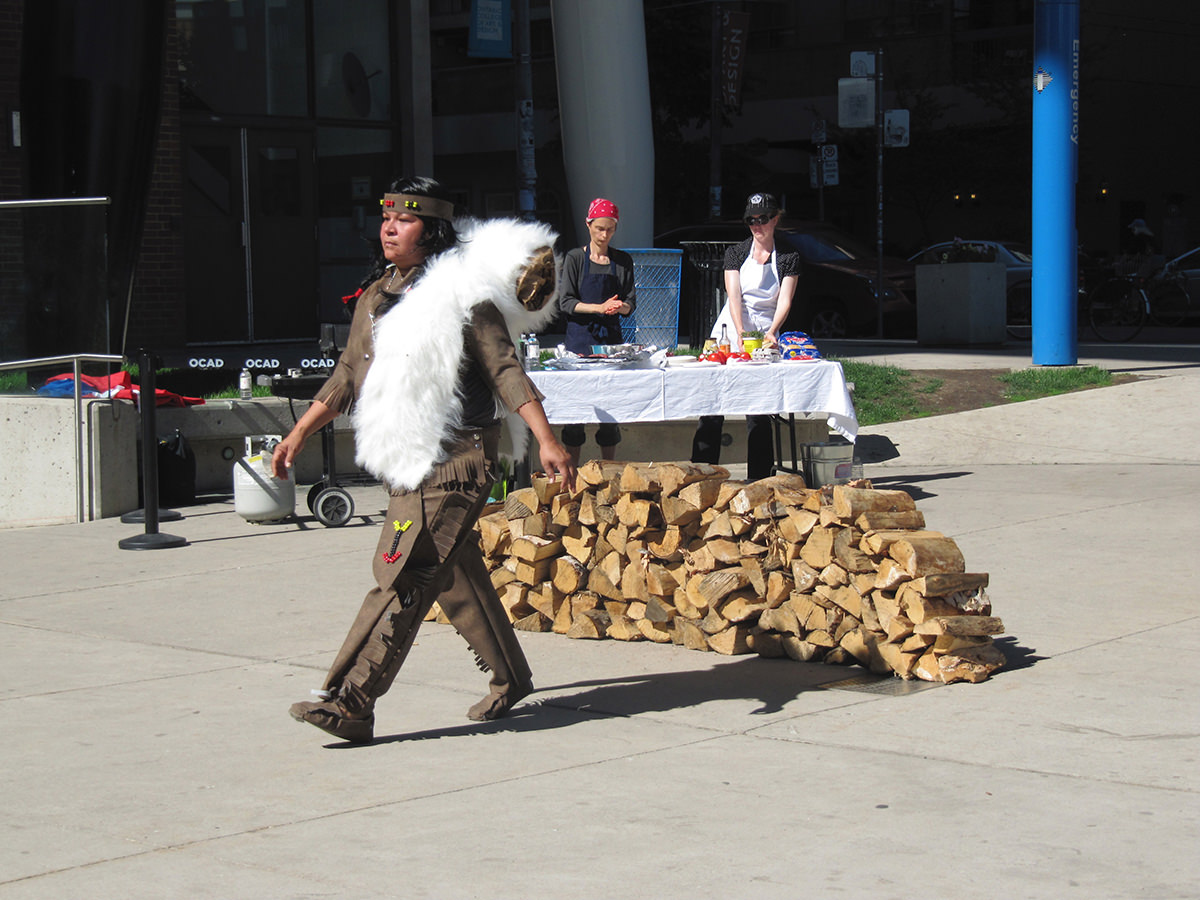 A woman in traditional Indigenous dress walks past a table manned by two people behind a stack of firewood. 