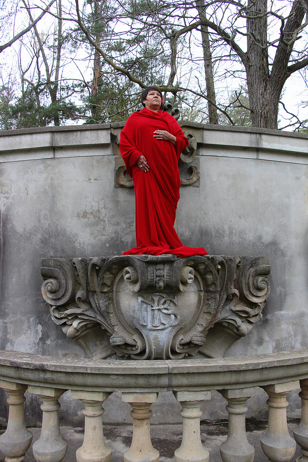 A woman wrapped in red cloth stands like a statue on a concrete monument. 