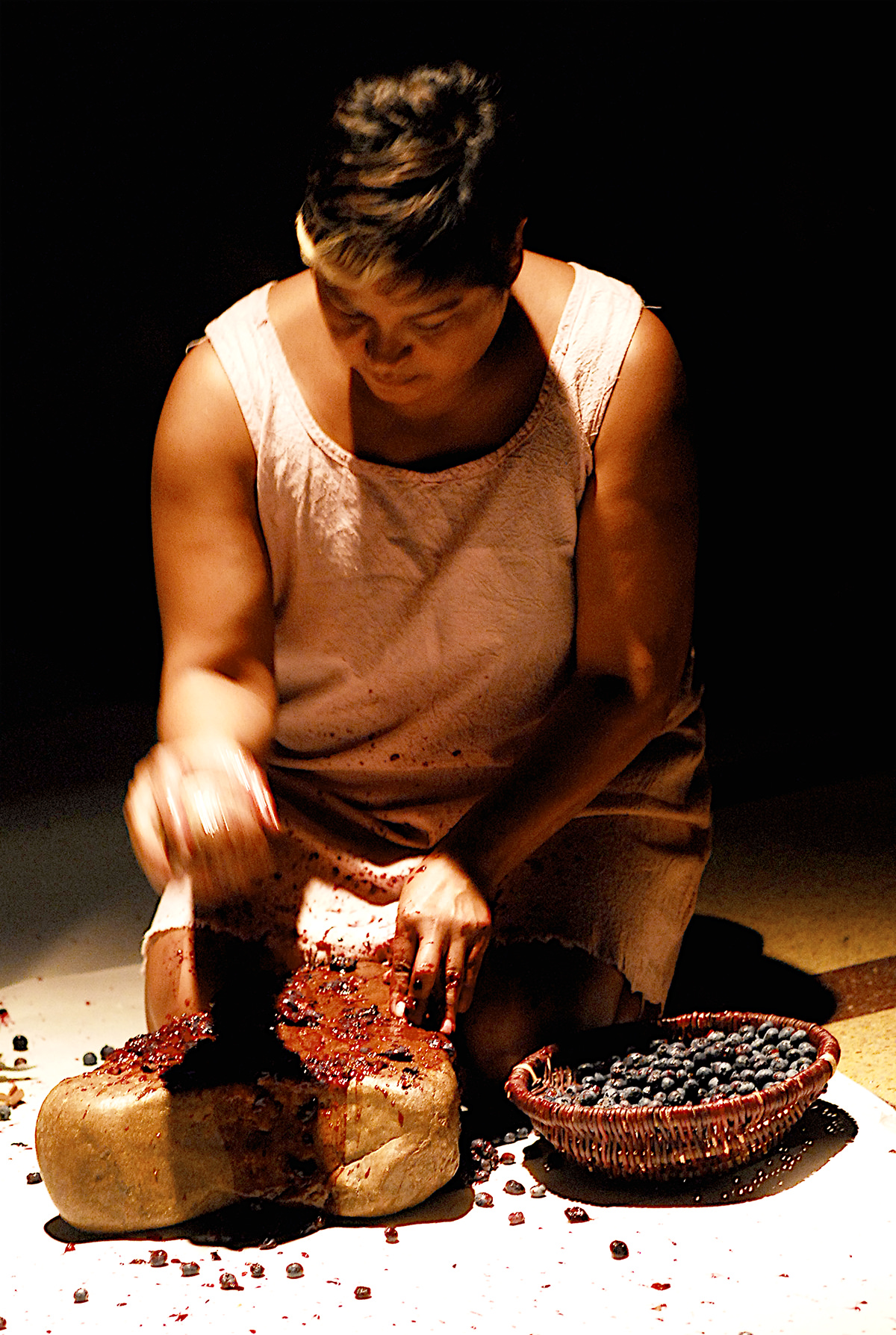 A woman kneels under a spotlight to work on an object with small fruit sitting in a bowl beside her. 