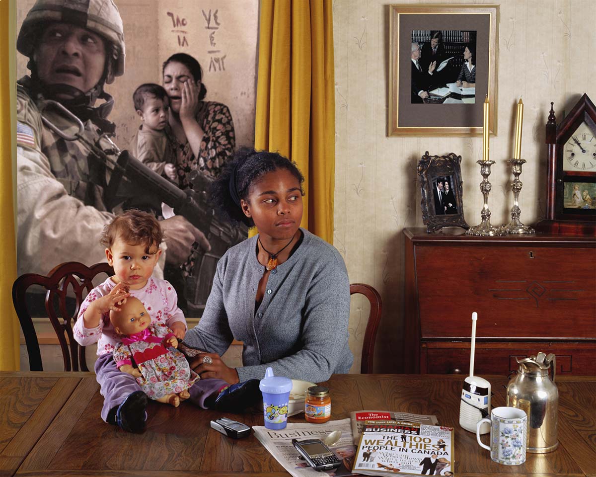 A woman sitting at a table with a child, with a war-themed photo in the background. 
