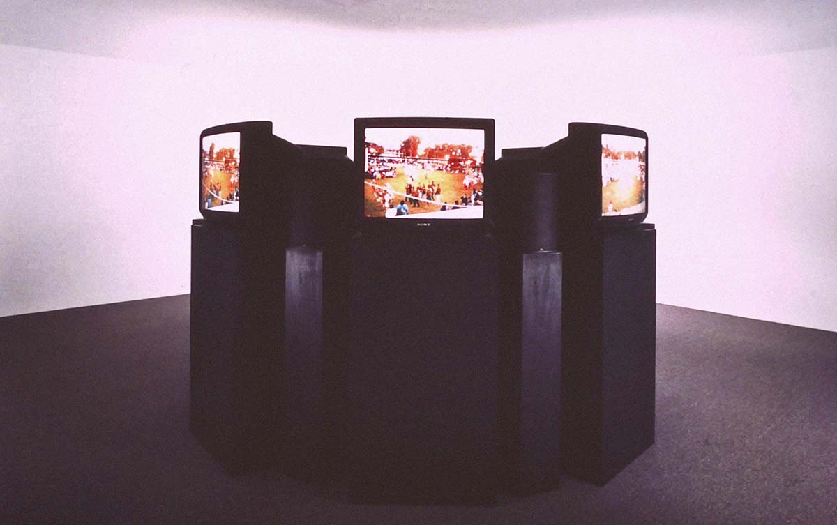 An installation showing several outward-facing television screens on black supports in a circle. 