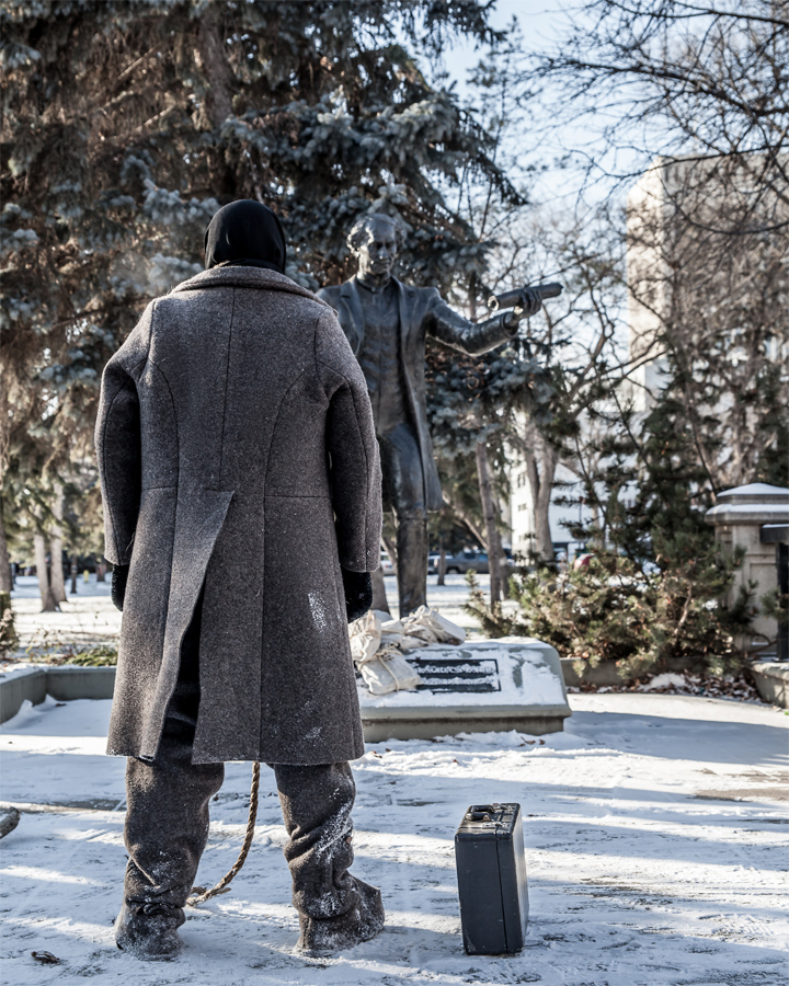 Winter scene showing the rear view of a stocky person wearing a black tuque and a felt coat. The person is standing in a park looking at a bronze statue of John A. Macdonald with an attaché case at their feet.  