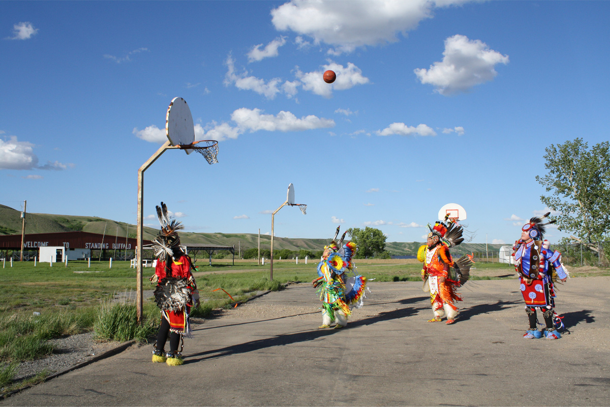 Four people wearing feathered headdresses and colourful, traditional regalia are seen shooting a basketball on an outdoor basketball court. 