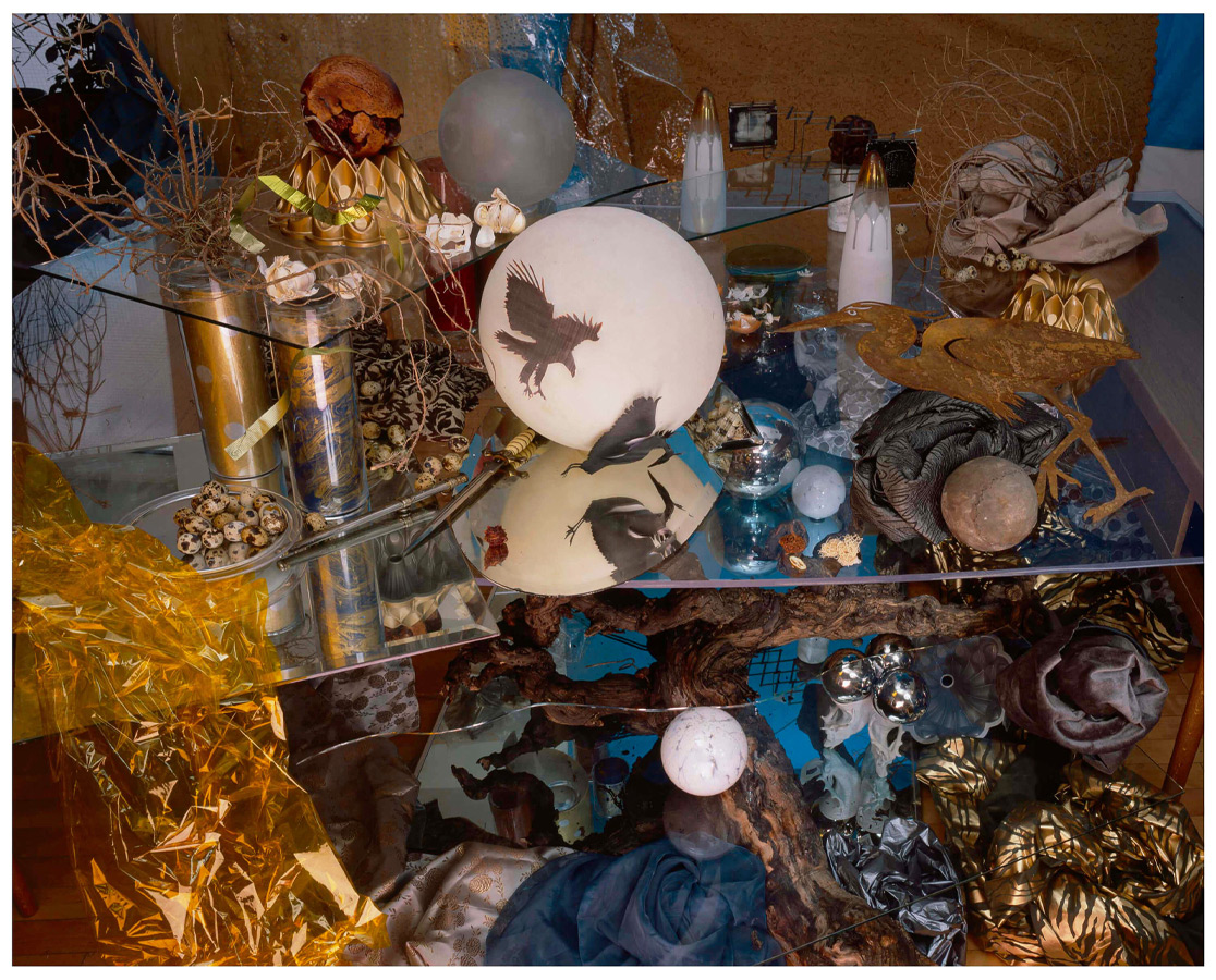 A complex array of orbs, small branches, metallic fabrics and bird images, on transparent shelves. Bronze and blue dominate the palette. 
