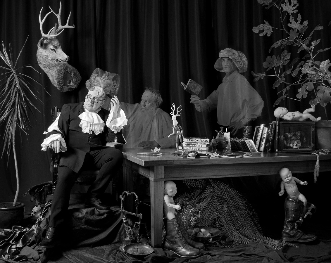 A man in dated garb sits beside a table. Ghosted figures, carrying ramshead masks, are behind. Figurines of infants are among other articles. 