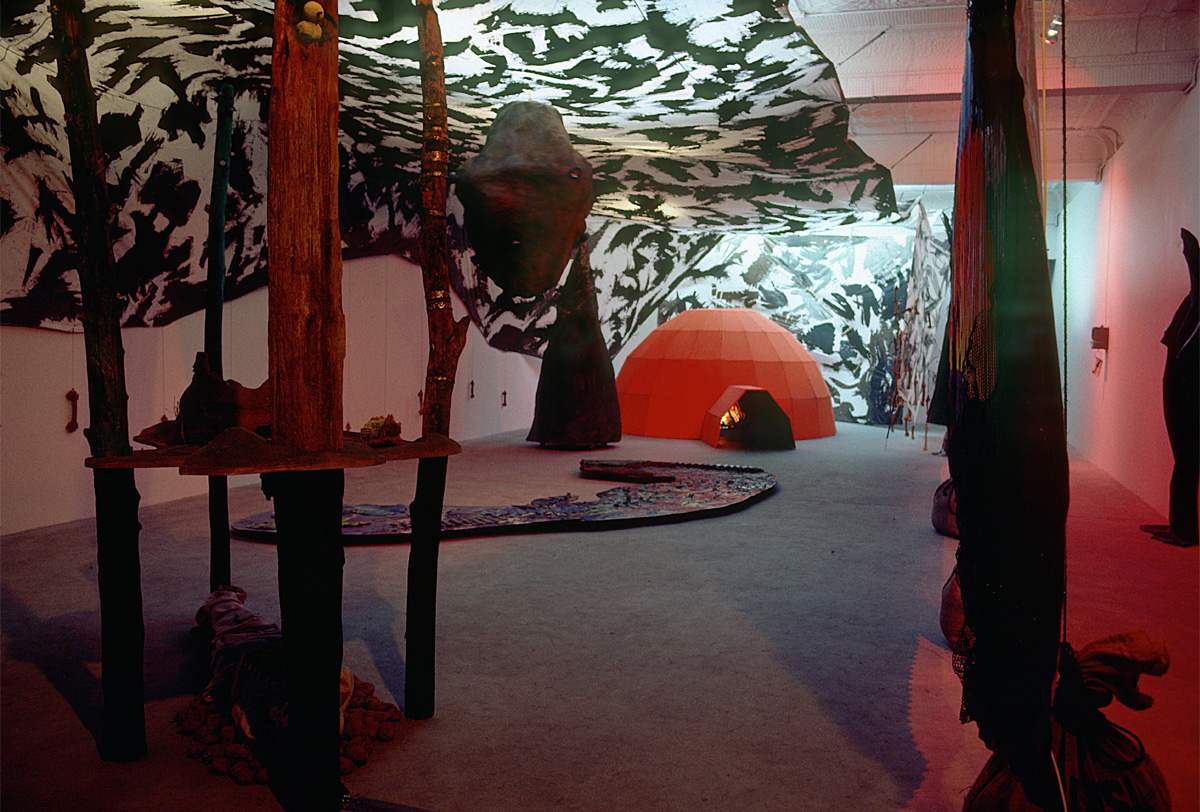 A white and black canopy is suspended above an orange igloo shape, several slender tree trunks and other objects in an arboreal theme. 