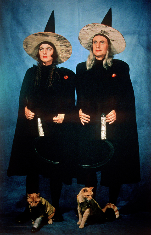 A portrait of two people in black robes and witch hats, carrying scythes. They stand against a blue backdrop. At their feet are two orange cats.  