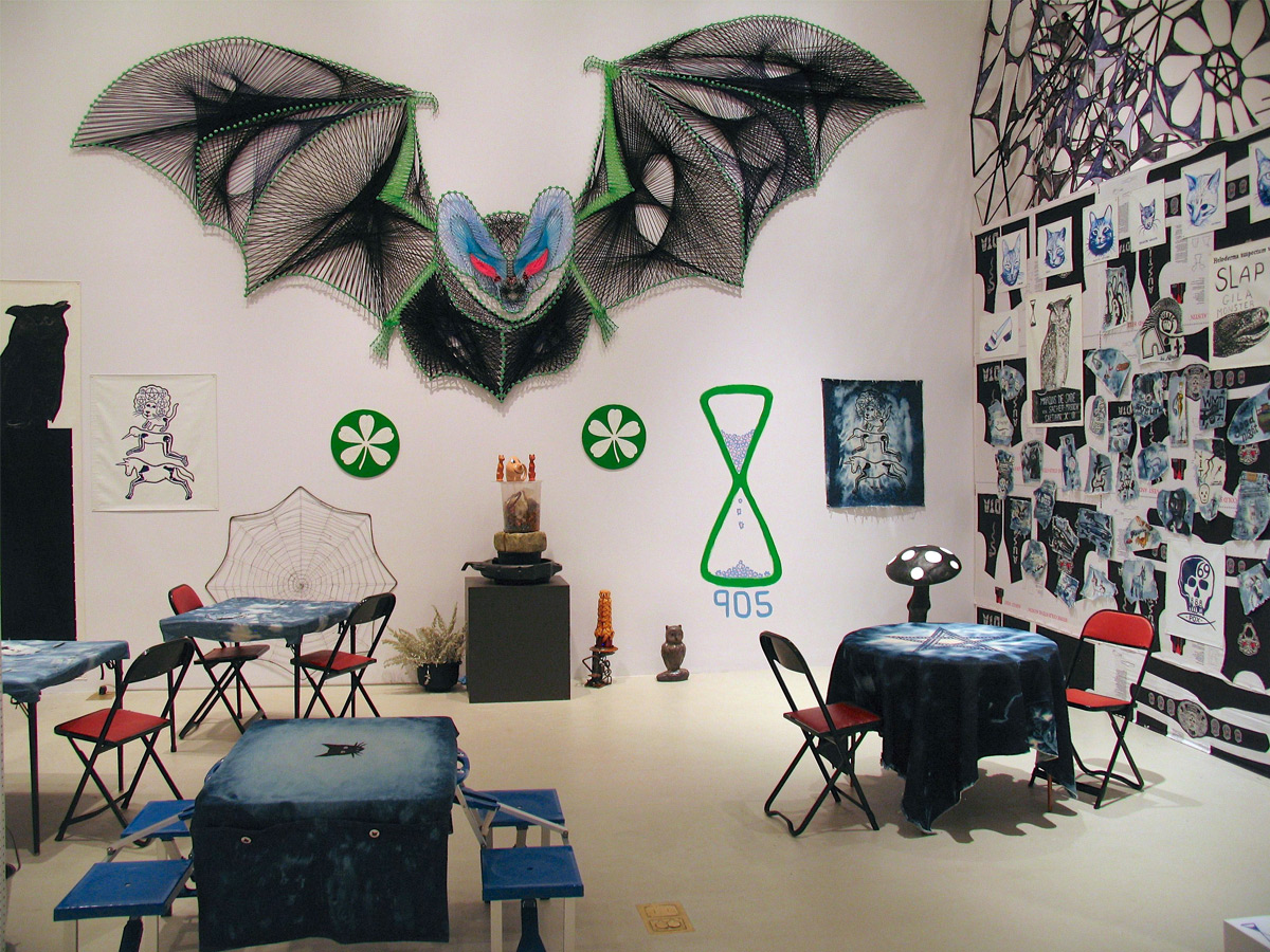 The form of a flying bat with red eyes dominates the space. On the walls are works depicting cats, skulls, logos, hourglasses and text. 