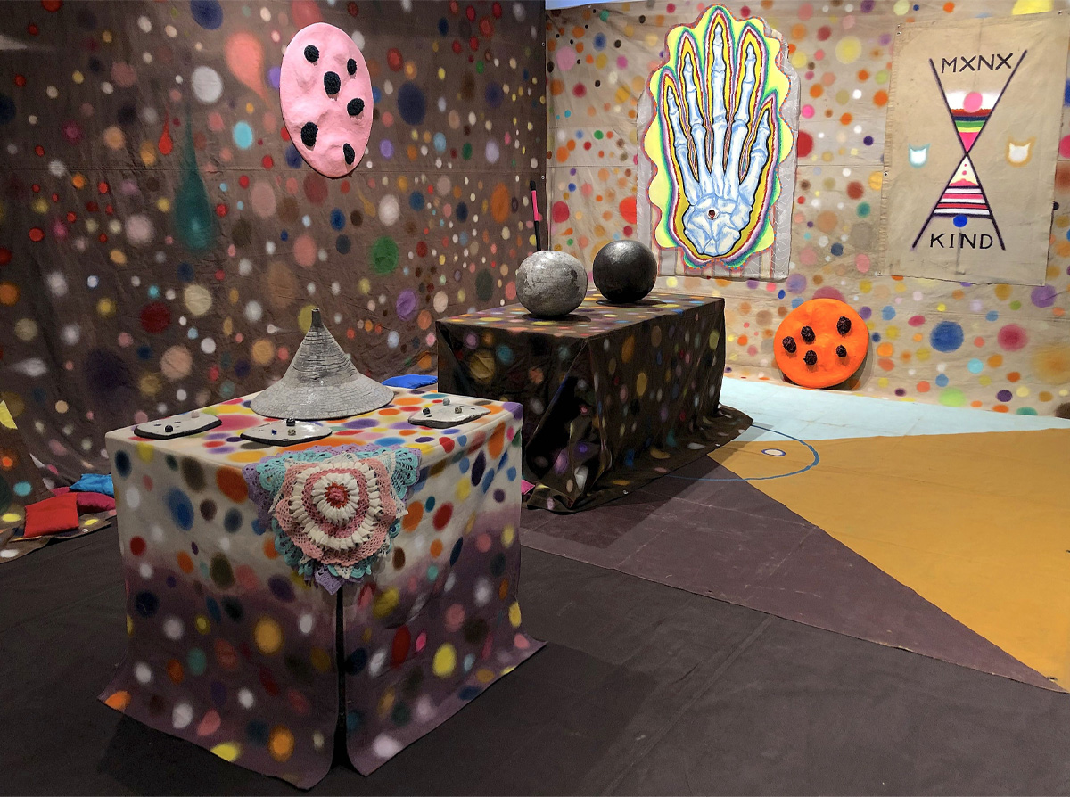 Tarpaulins painted with coloured circles cover walls and two tables. Among objects on them are grey spheres and a painting of a skeletal hand.  