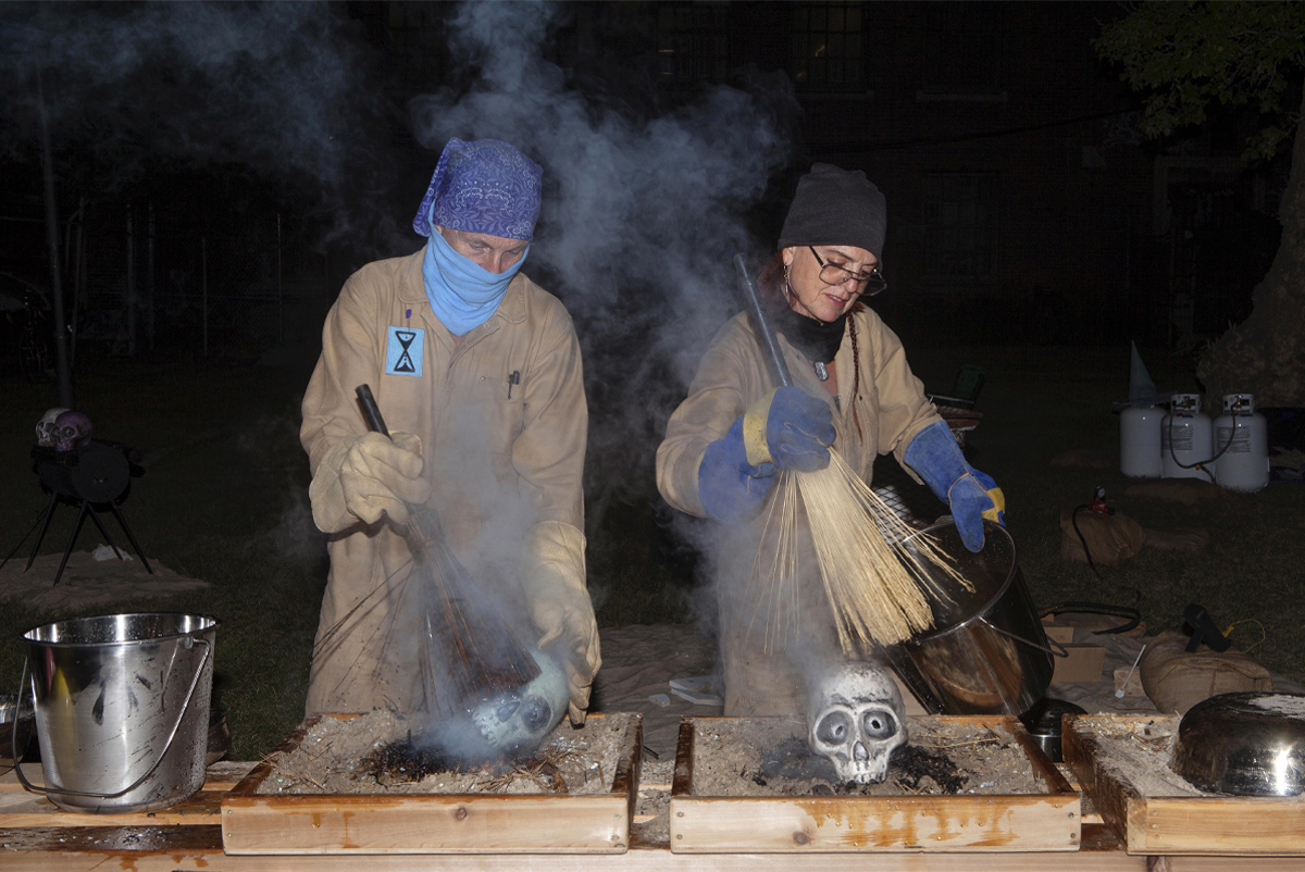 Outdoors at night, two people in coveralls and gloves hold buckets and daub ceramic skulls with whisk brooms. Smoke rises above their heads. 