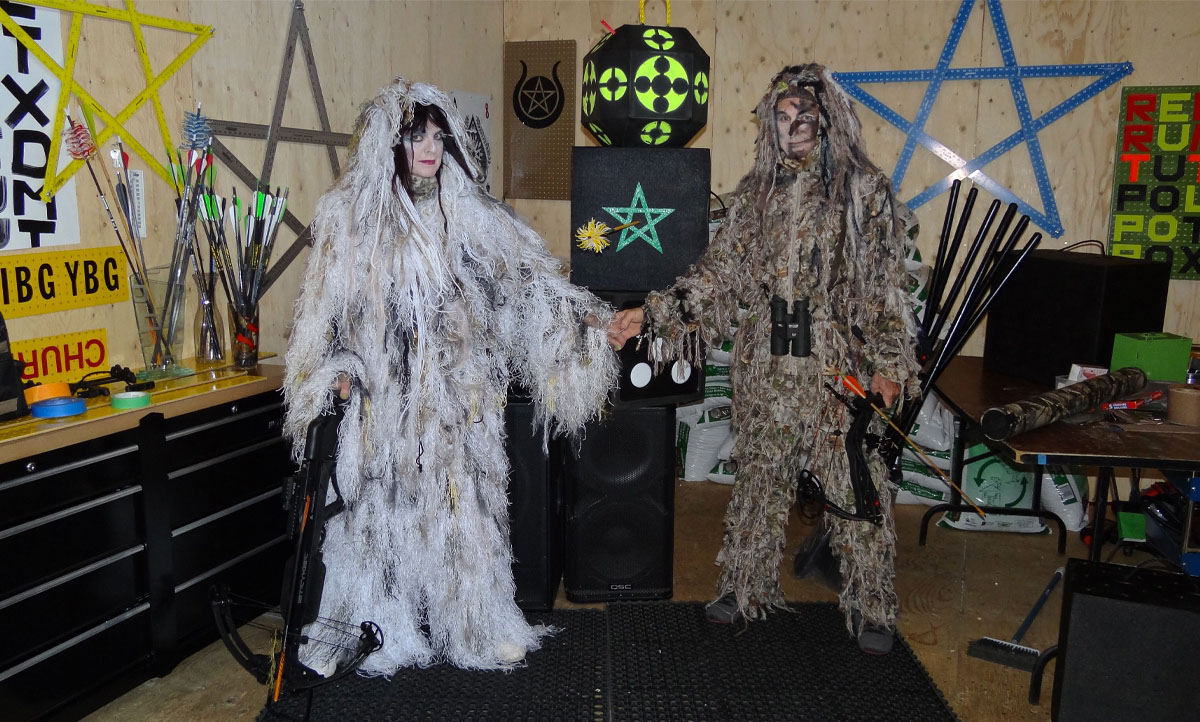 The photograph shows two human figures in furry suits holding hands. Each carries a crossbow. Walls bear various pentagrams and lettering. 