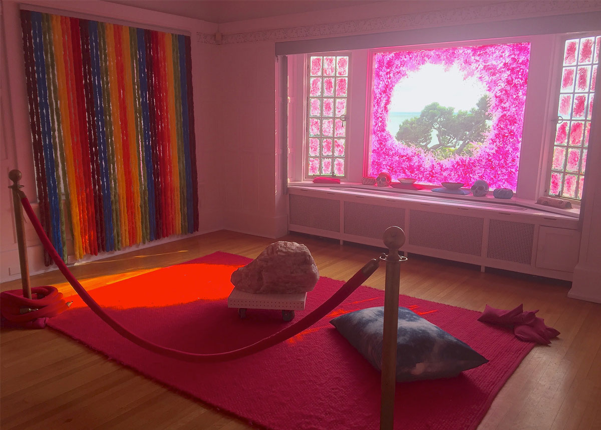 A large rock and a pillow rest on a red rug. A window is daubed with pink. Hanging fabric tubes form a large rainbow curtain on a wall.  