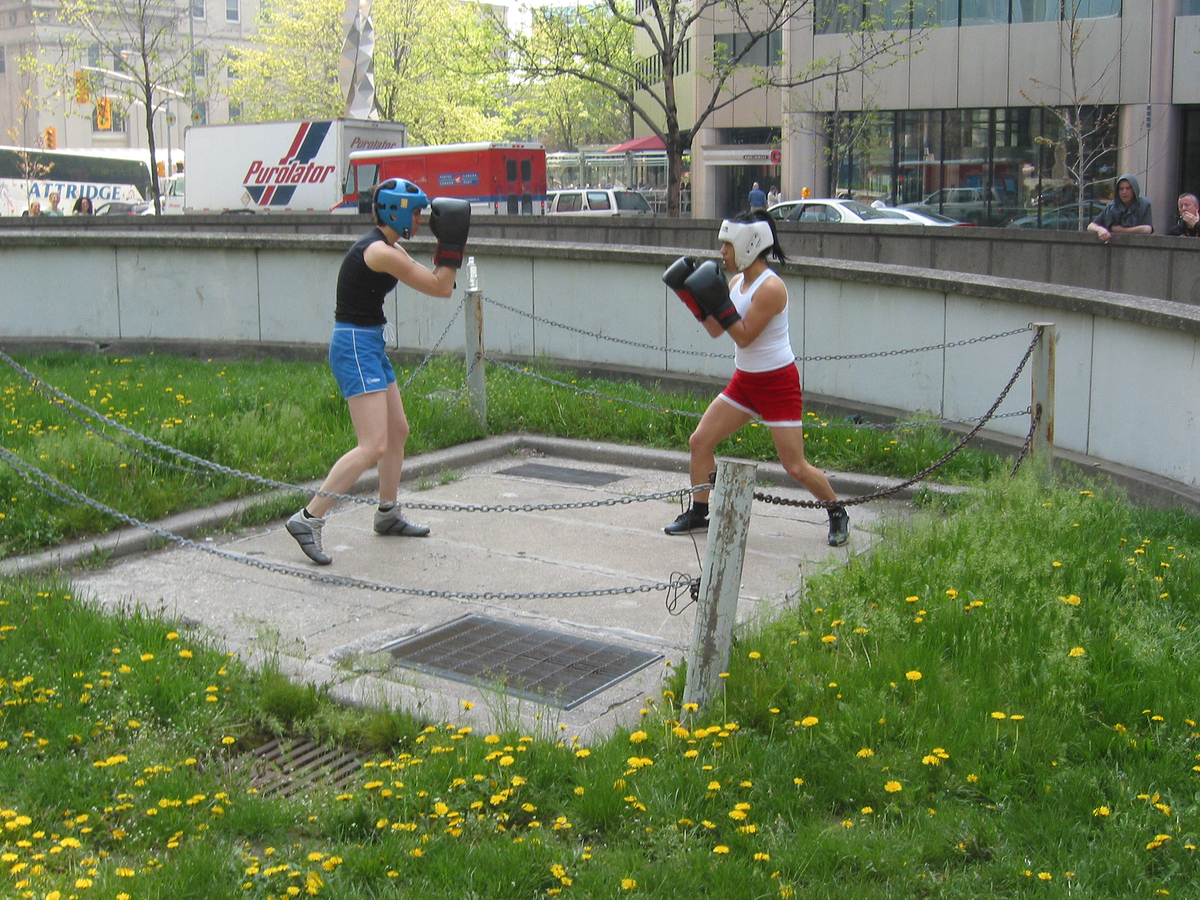 Two young people wearing boxing gloves and helmets engage in boxing while standing on a concrete pad near a ramp to underground parking. 