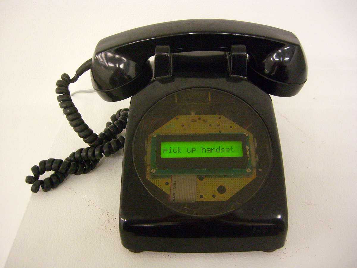 A black, 1970s-era telephone sits on a white surface. Its face shows a green LED display illuminated with the words "Pick up handset." 