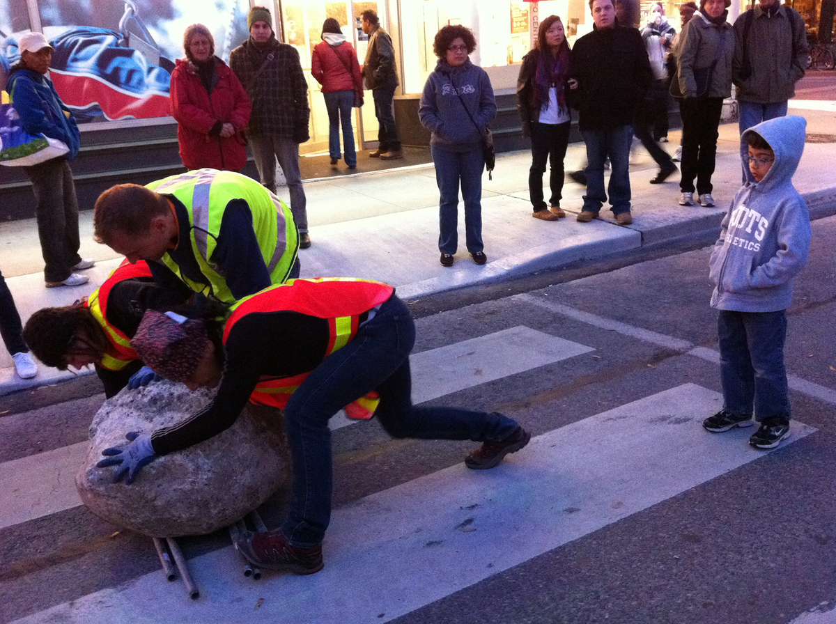 Three men in reflective vests strain to move a large boulder lying on the pavement of a city street as bystanders watch. 
