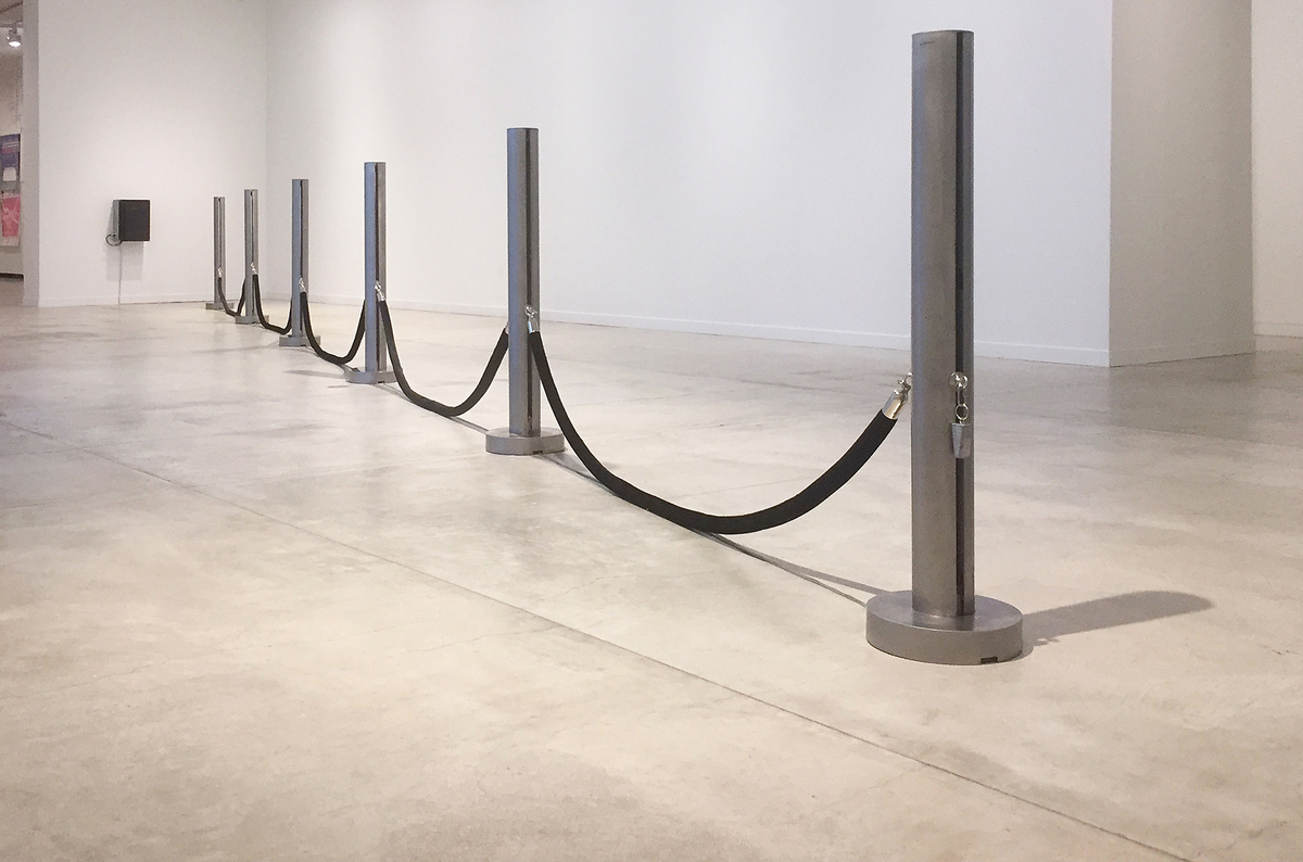 Sections of black tubes hang between equally spaced metal stanchions that sit in a row on a gallery floor. A black box is attached to a wall. 