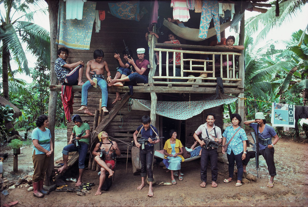 Men, women and children pose on two levels of a wooden dwelling in a jungle setting. Some of them hold guns. 