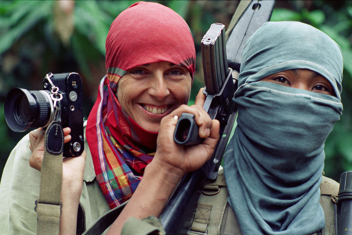 The artist, in a red headscarf, smiles as she holds a camera beside her head. In front, a man in a balaclava holds an assault rifle. 