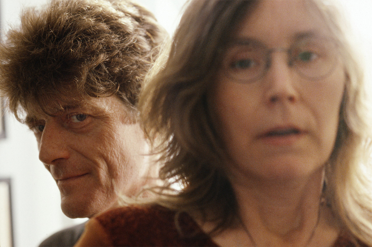 A closeup shows a man's face as he looks over the shoulder of a woman. Both are facing the viewer. 