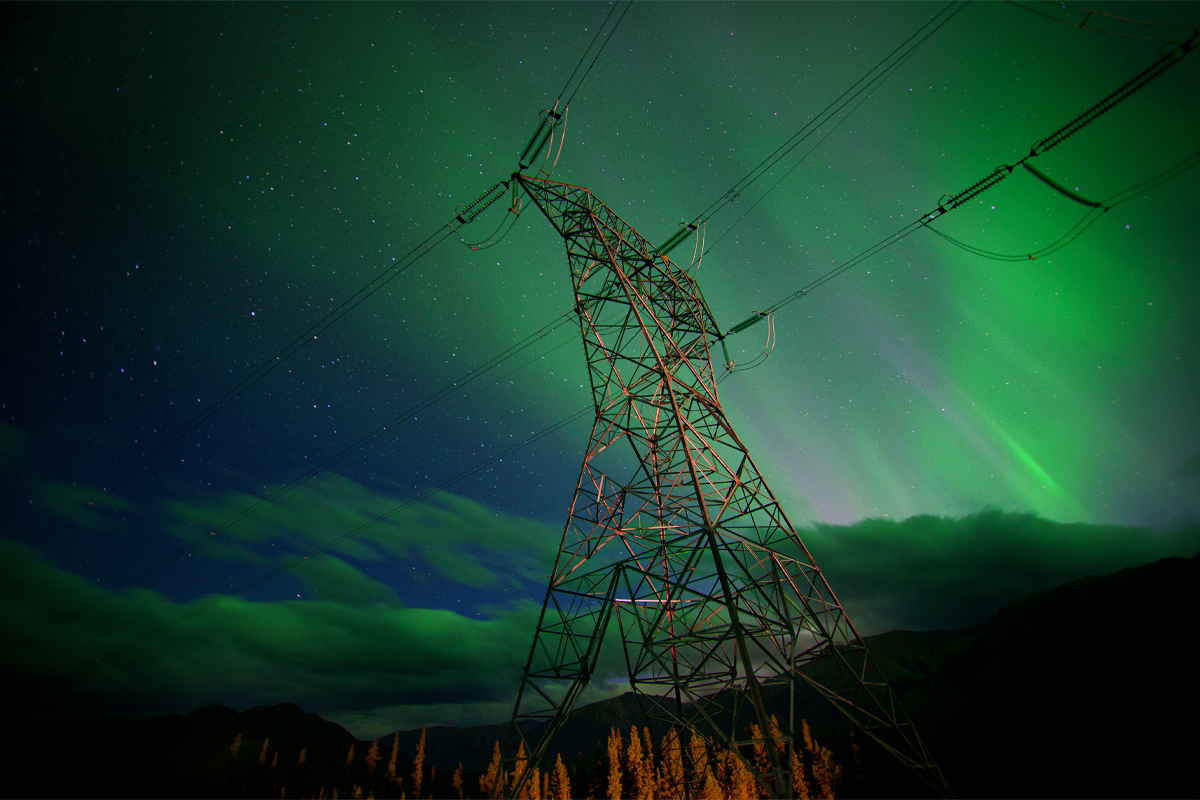 A steel hydro tower is seen against a backdrop of bright green northern lights and a starry sky. The horizon is a row of dark mountains. 
