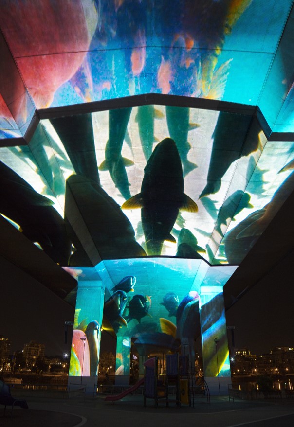 Images of swimming fish are projected onto the bottom surface of a concrete bridge at night, creating the impression of an overhead aquarium. 