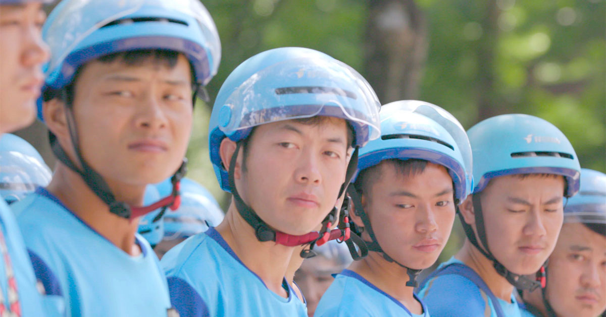 Young men stand shoulder to shoulder, wearing light blue helmets with visors and matching jerseys. They appear to be apprehensive. 