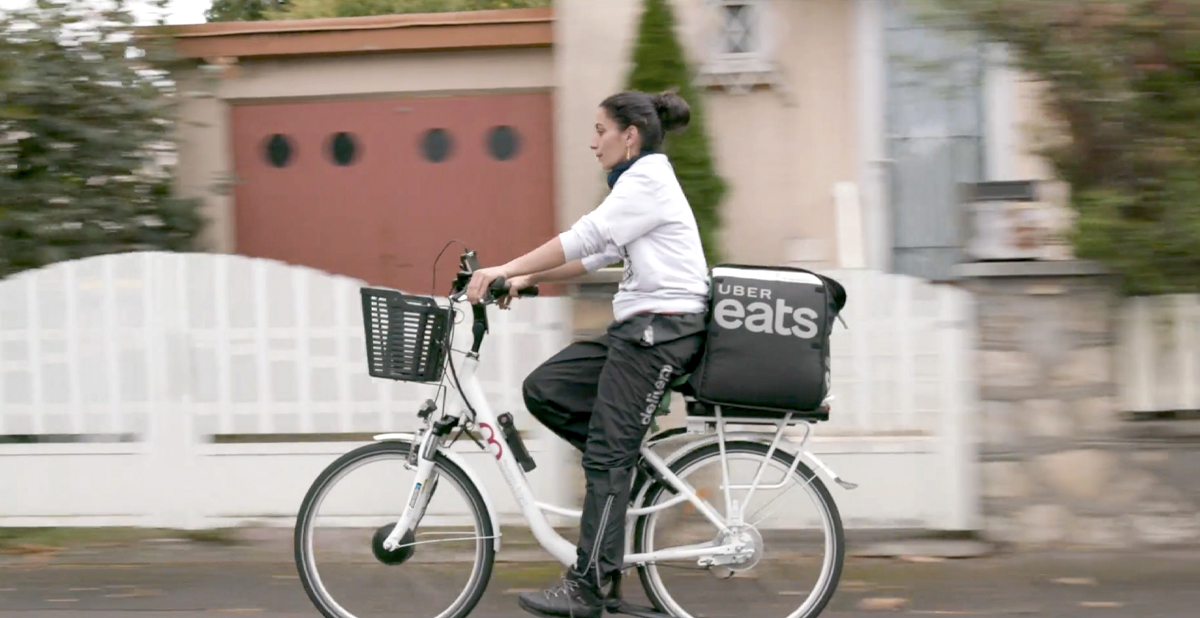A woman rides a bicycle with an "Uber Eats" carrier past a gated house. She wears earrings, rain pants and a long-sleeved pullover shirt.  