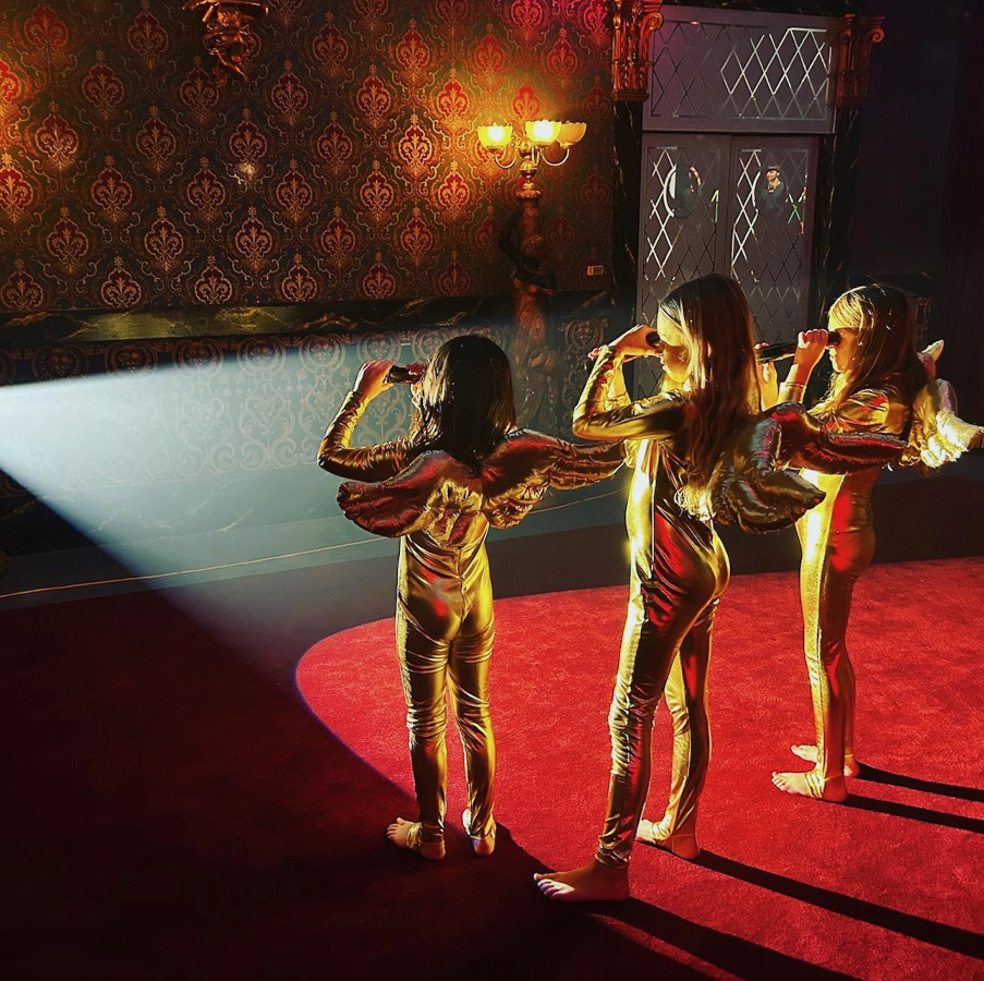 Three young girls stand barefoot in a spotlight on a red carpet. They wear gold suits with wings and hold small telescopes up to their eyes. 