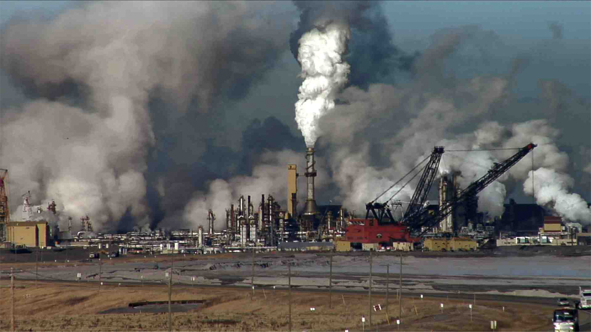 The sky is filled with smoke and steam rising from the silver stacks of a refining plant. Large cranes sit at the edge of a body of water. 