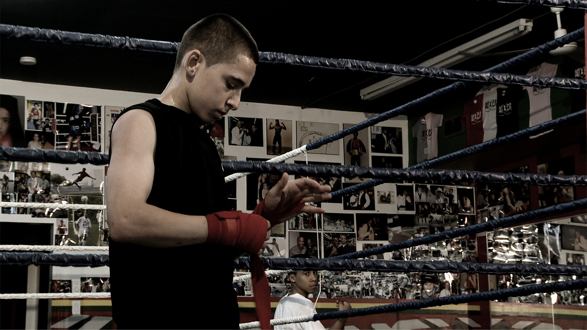 A young man in front of a boxing ring wraps his hands. The walls behind him are hung with t-shirts and photographs of athletes. 