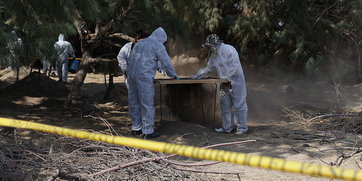 People in white hazmat suits work while standing at a table under trees. Caution tape is stretched across the foreground. 