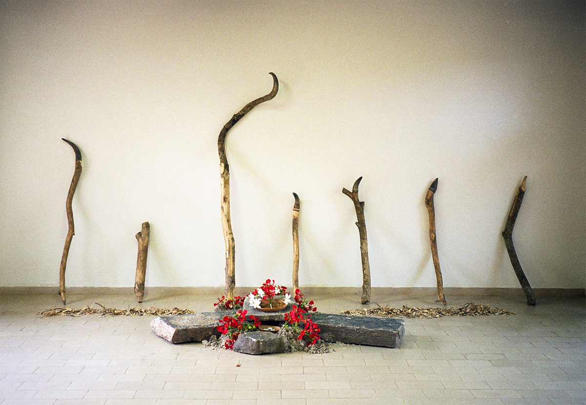 Seven wooden sticks, some capped with horns, lean against a wall. In front are slabs of stone topped by flowers, wooden bowls and candles. 