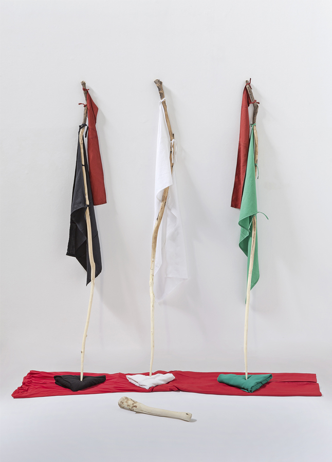 Pieces of white, black, red and green cloth, tied to three sticks leaning on a white wall. They rest on pieces of fabric in similar colours. 