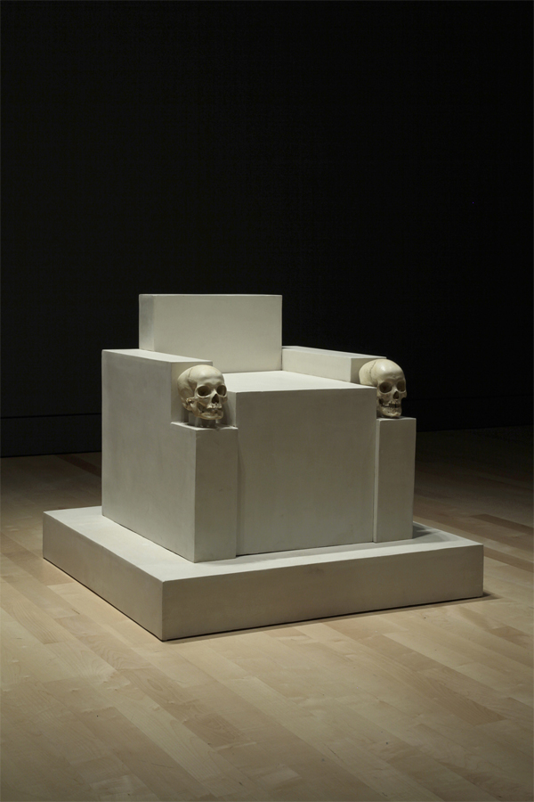 Boxy white shapes form an austere, low throne. A human skull fronts each armrest. It rests on a matching plinth. 