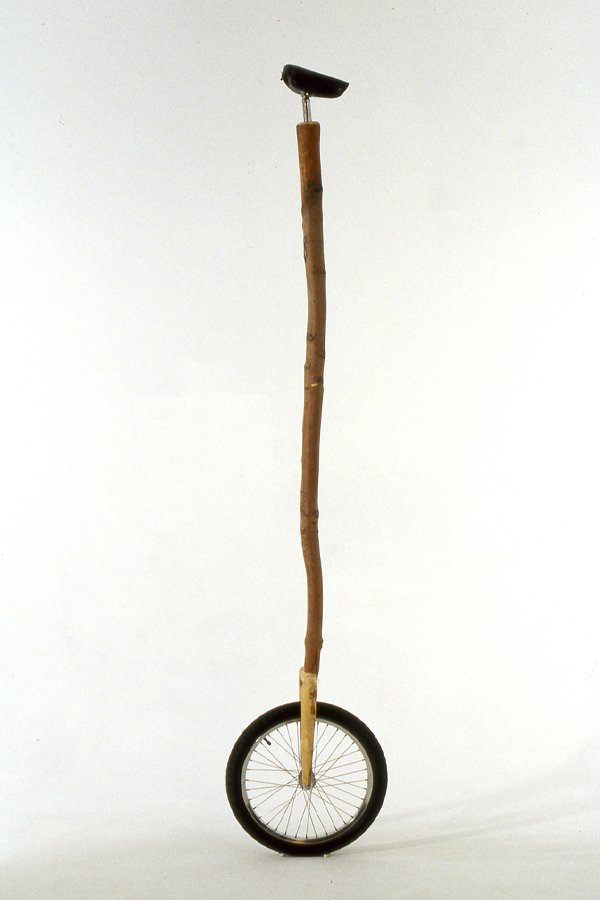 A long wooden staff rises from the axle of a small bicycle wheel. It is topped by a small black seat. 