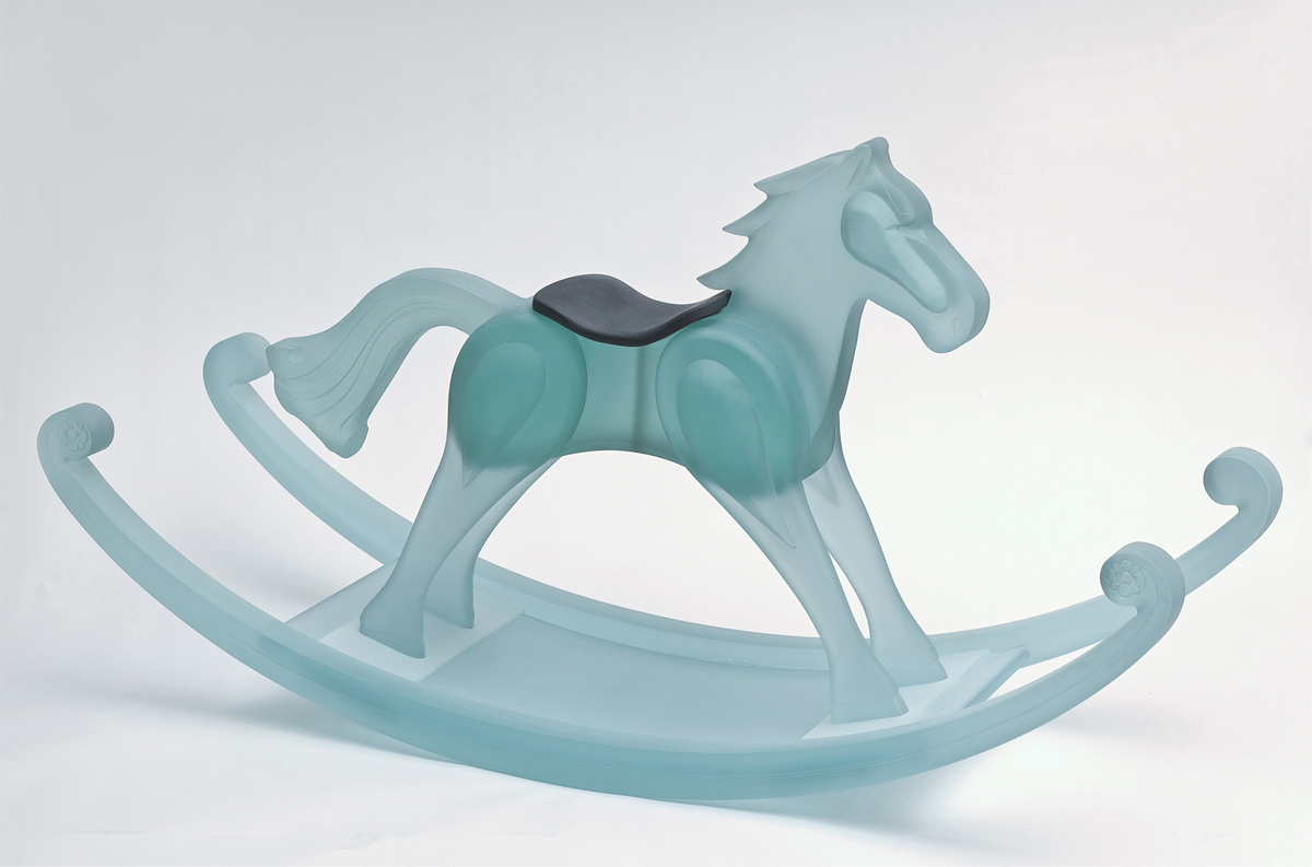 A life-sized rocking horse is formed of frosted translucent blue-green glass. In contrast, the horse wears a black saddle. 