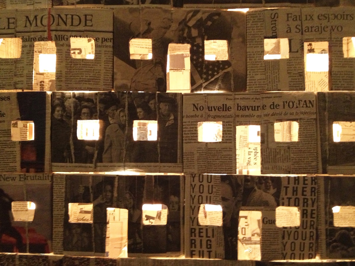 A structure covered in newspaper articles with rectangle cutouts letting out light from within.  