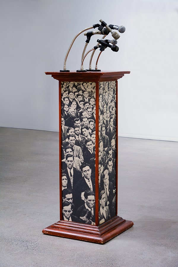A wooden lectern covered with pictures of faces of mouthless men. 