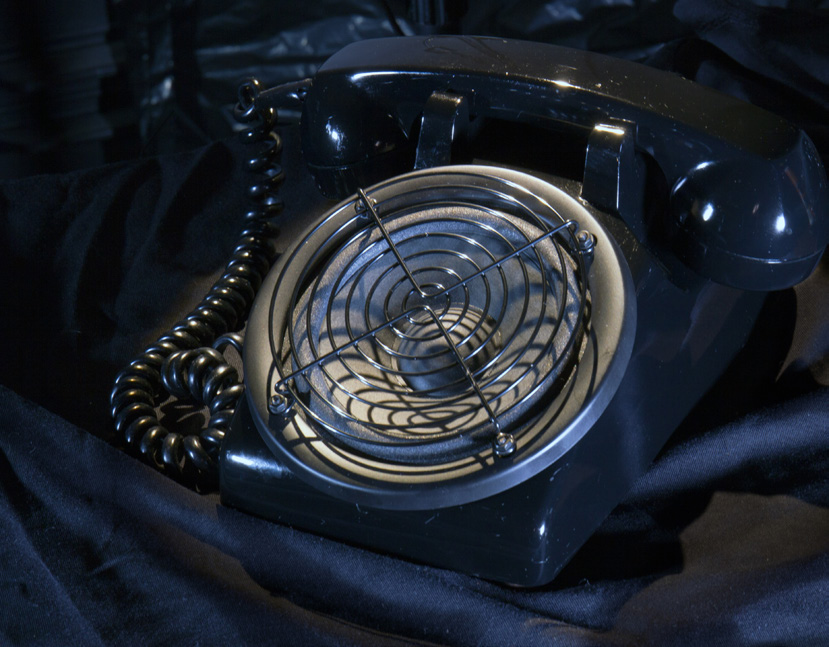 A black rotary phone whose dial has been replaced by a speaker with a grille sits on black fabric. 