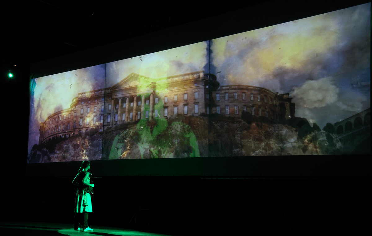 A person watches a projection of herself superimposed onto a picture of a historical building. 